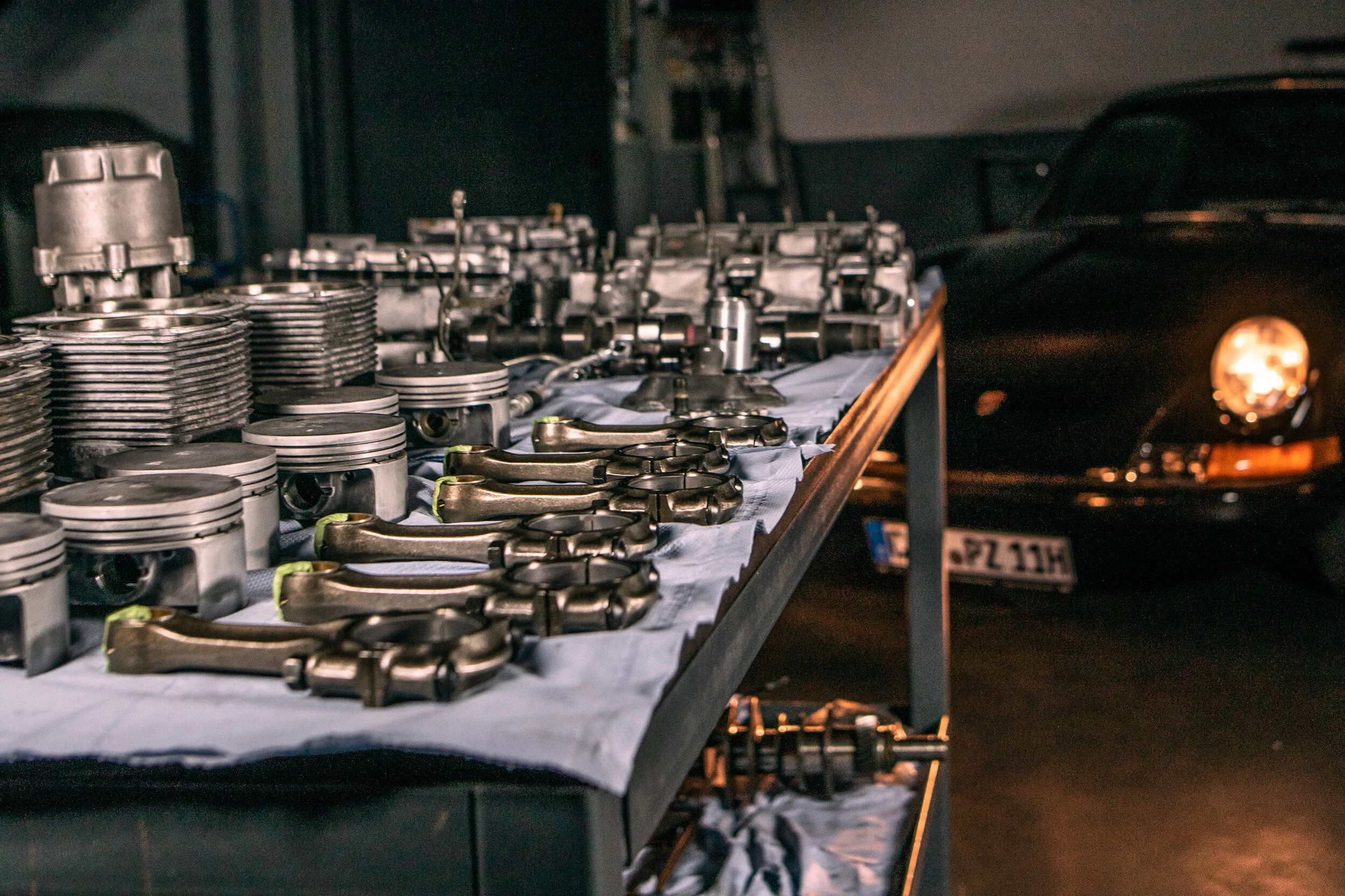 Kolben, Pleuel und Zylinder eines zerlegten Boxermotors auf der Werkbank, dahinter ein Porsche 911